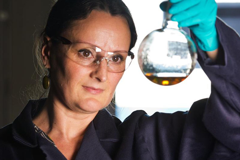 A chemist wearing a Chevron lab coat and protective gear checks the chemical inside a flask by holding it up to the light. 